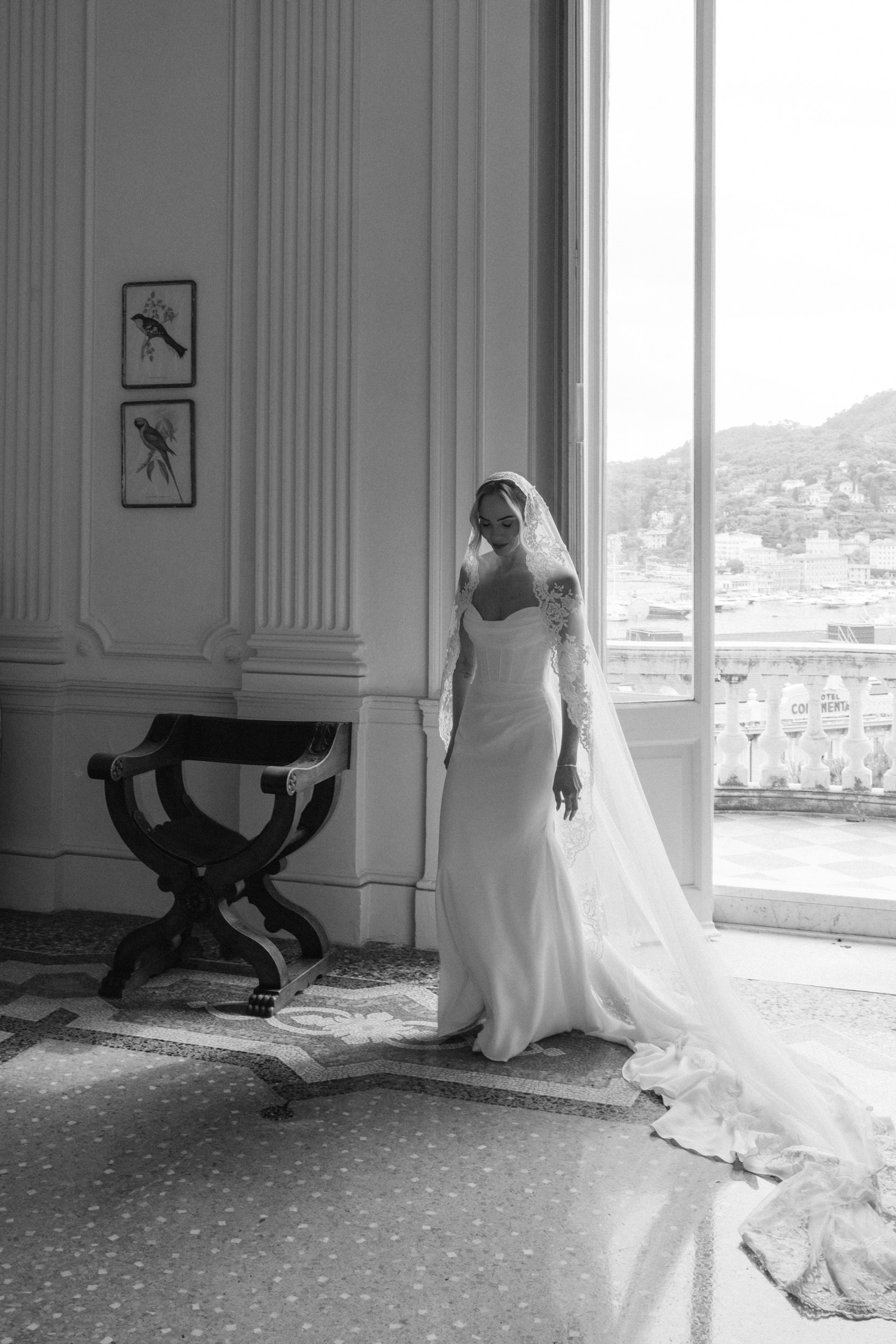 Black and white photo of bride in strapless gown and cathedral veil standing by a tall window overlooking Santa Margherita Ligure