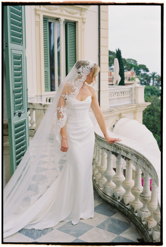 Film photograph of bride from behind in lace-edged veil, leaning on a balcony railing overlooking the Italian coastline