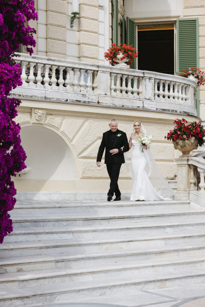 Father of the bride walking bride down marble steps past bougainvillea, bride carrying white bouquet
