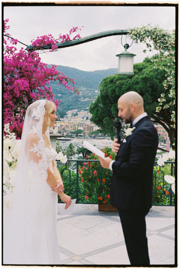 Film photograph of bride and groom facing each other during outdoor vow exchange, coastal town visible below