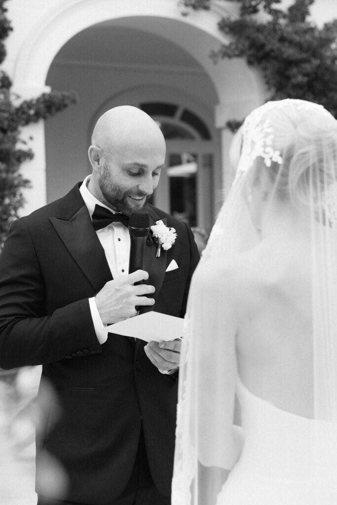 Black and white photo of groom reading vows into a microphone, bride's veil in the foreground