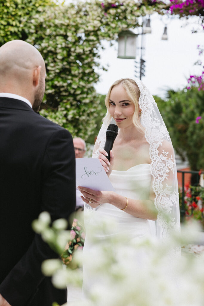 Bride reading vows into a microphone, card reading "her" in hand, groom listening