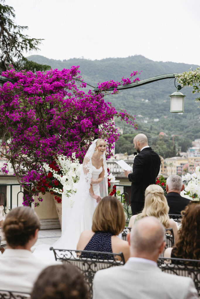 Bride laughing during vow exchange under a bougainvillea arch, groom reading from a card, guests seated below