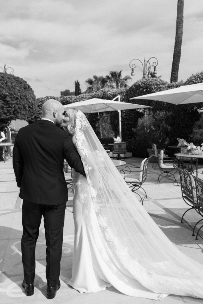 Black and white photo of bride and groom kissing from behind, veil trailing across a hotel terrace