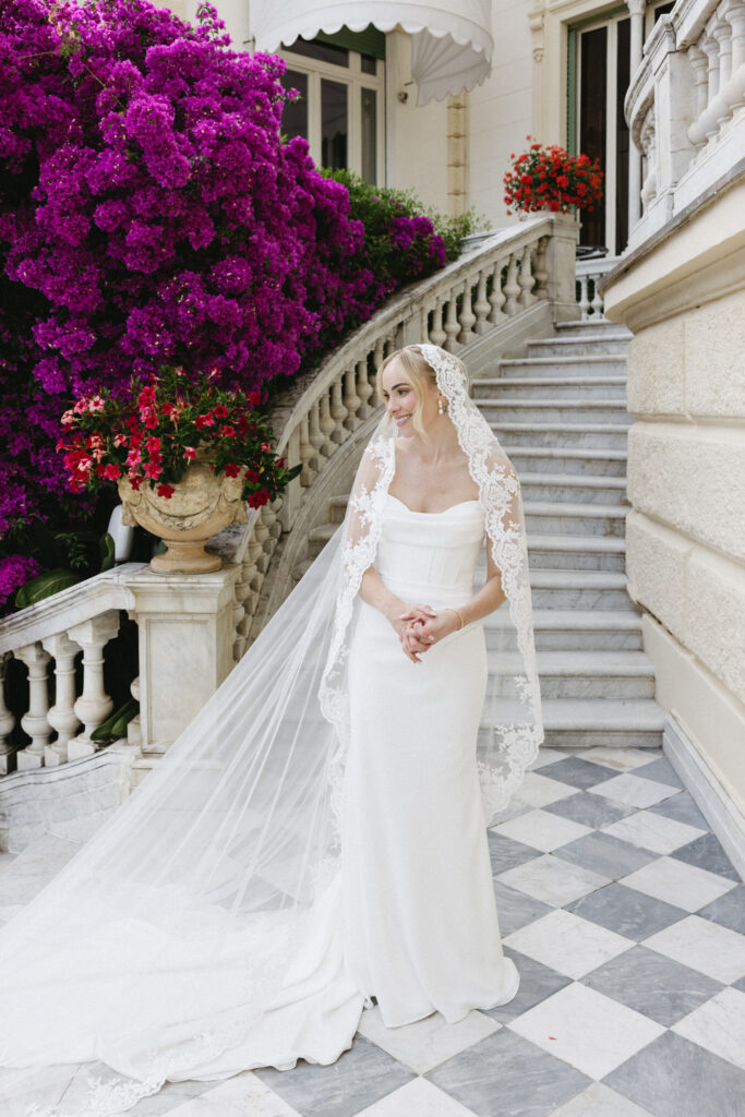 Bride in lace-edged veil smiling on marble steps beside bougainvillea, groom's hotel entrance in background
