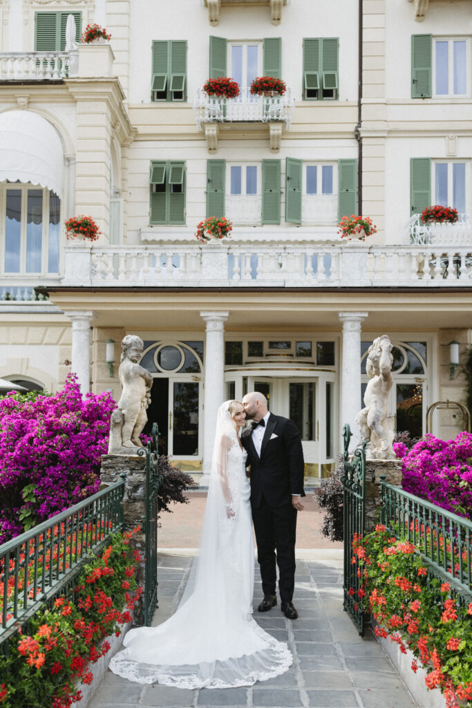 Bride and groom kissing on foreheads outside the hotel entrance, flanked by statues and bougainvillea