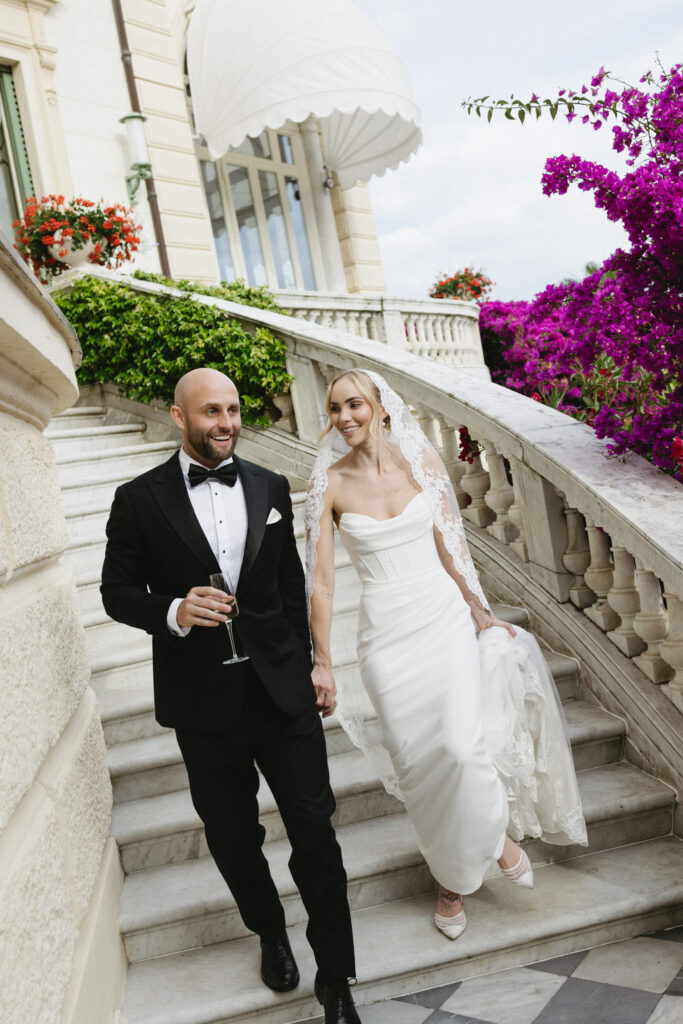 Groom and bride walking down marble steps holding champagne, bougainvillea cascading beside them
