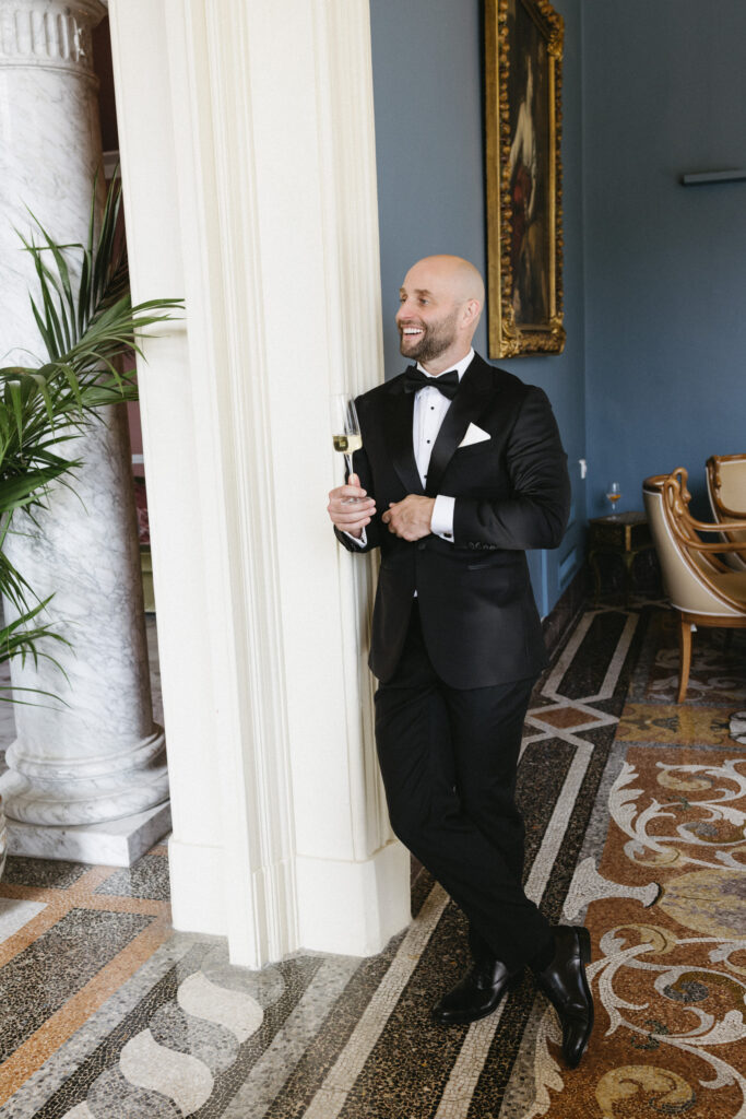 Groom in tuxedo laughing while leaning against a marble column, holding a champagne flute in an ornate interior room