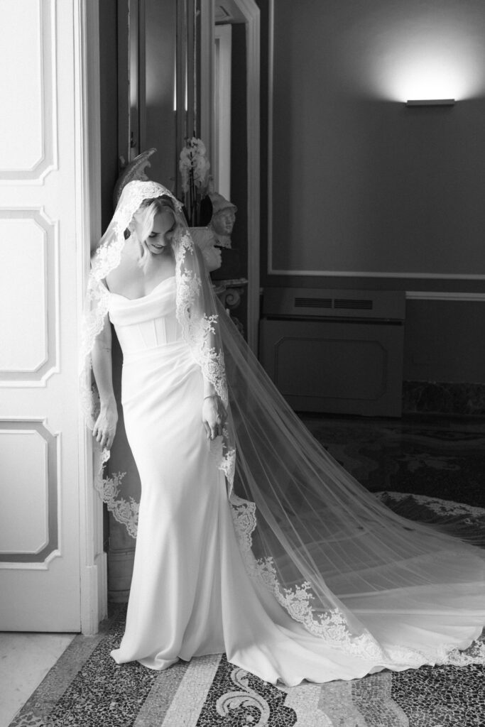 Black and white photo of bride in lace-edged veil standing in a doorway, veil trailing across a mosaic floor