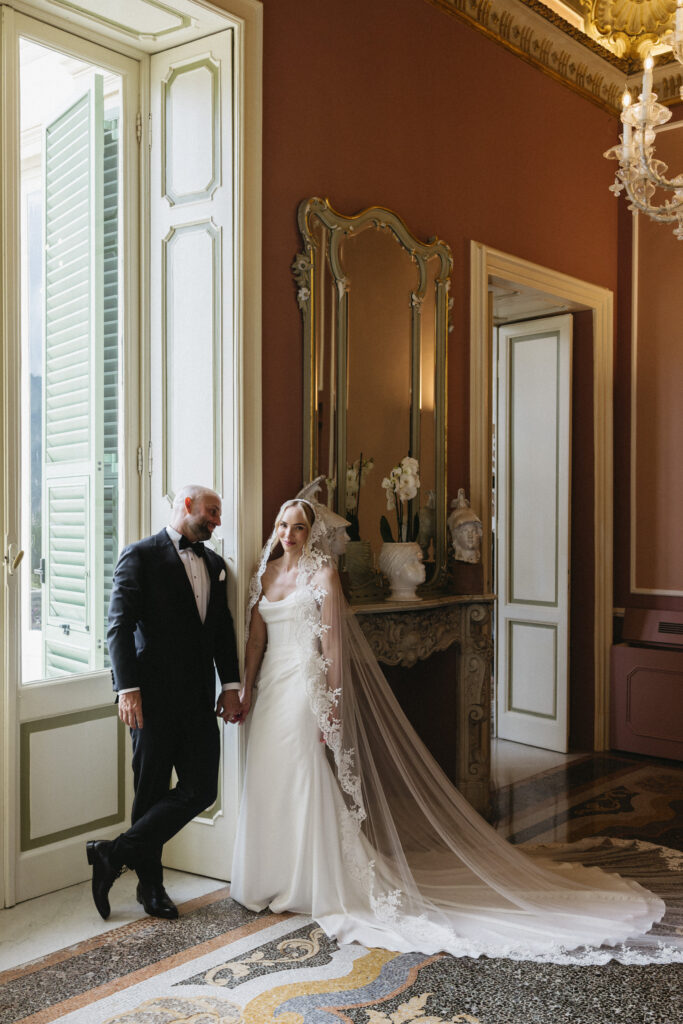 Bride and groom holding hands in an ornate hotel sitting room with a gilded mirror and marble fireplace, veil trailing across the mosaic floor