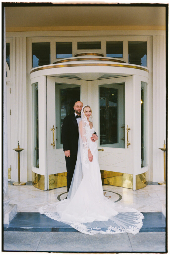 Film photograph of bride and groom standing under the hotel's revolving door canopy, veil pooling on the marble floor