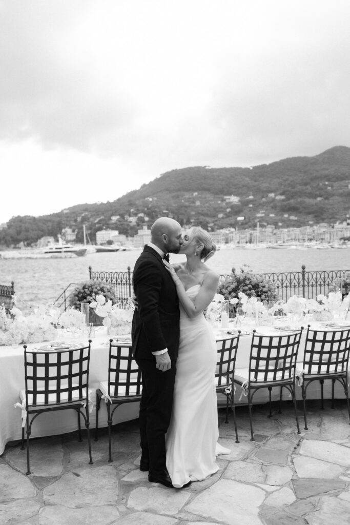 Black and white photo of bride and groom kissing beside their reception table set with white florals overlooking the sea