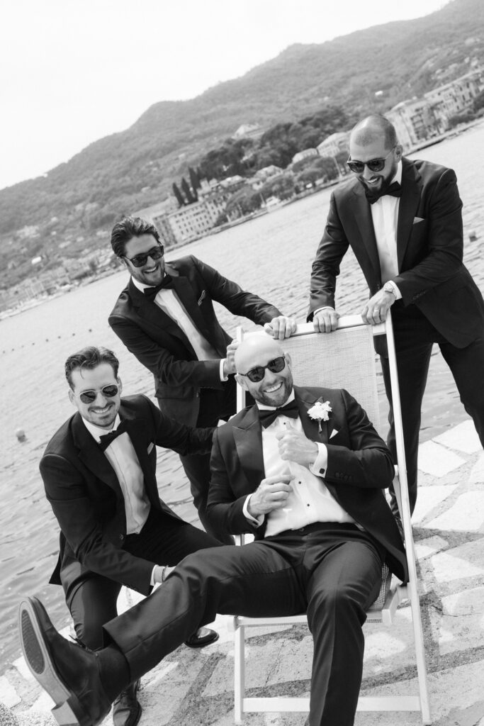 Black and white photo of groom seated in a deck chair surrounded by three laughing groomsmen on a waterfront terrace