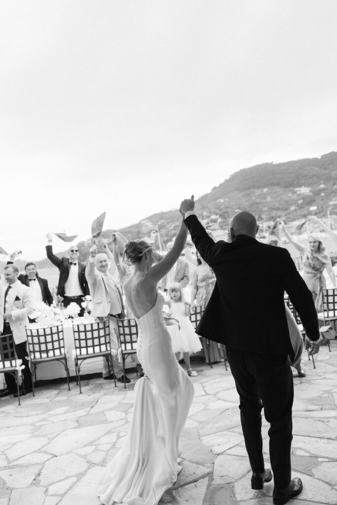 Black and white photo of bride and groom walking hand in hand toward cheering guests waving napkins at the reception