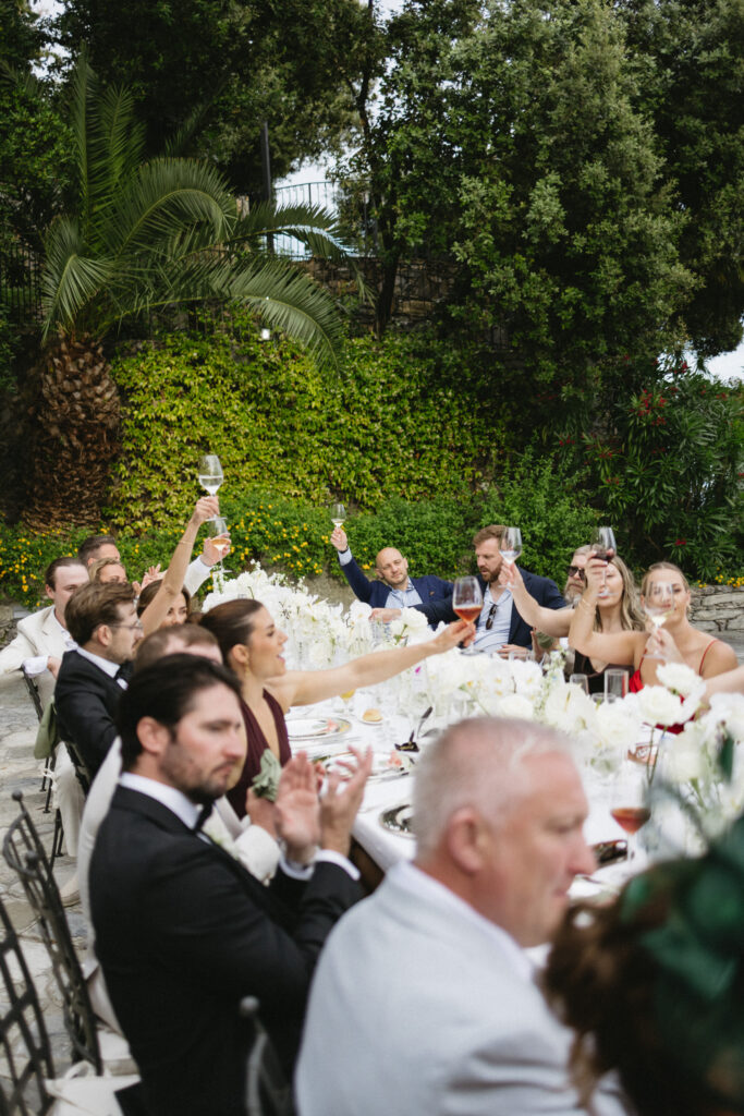 Reception guests raising wine glasses in a toast at a long outdoor table surrounded by greenery