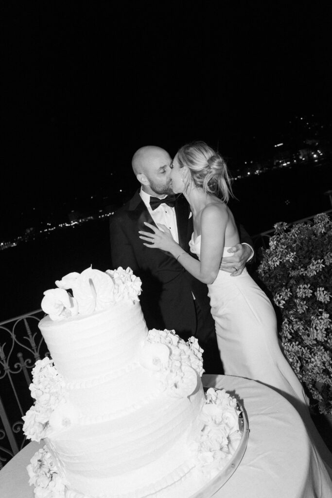 Black and white photo of groom biting into a tiered wedding cake decorated with white flowers, bride laughing beside him