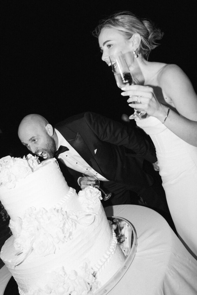 Black and white photo of bride laughing while holding a champagne glass, groom biting the wedding cake beside her