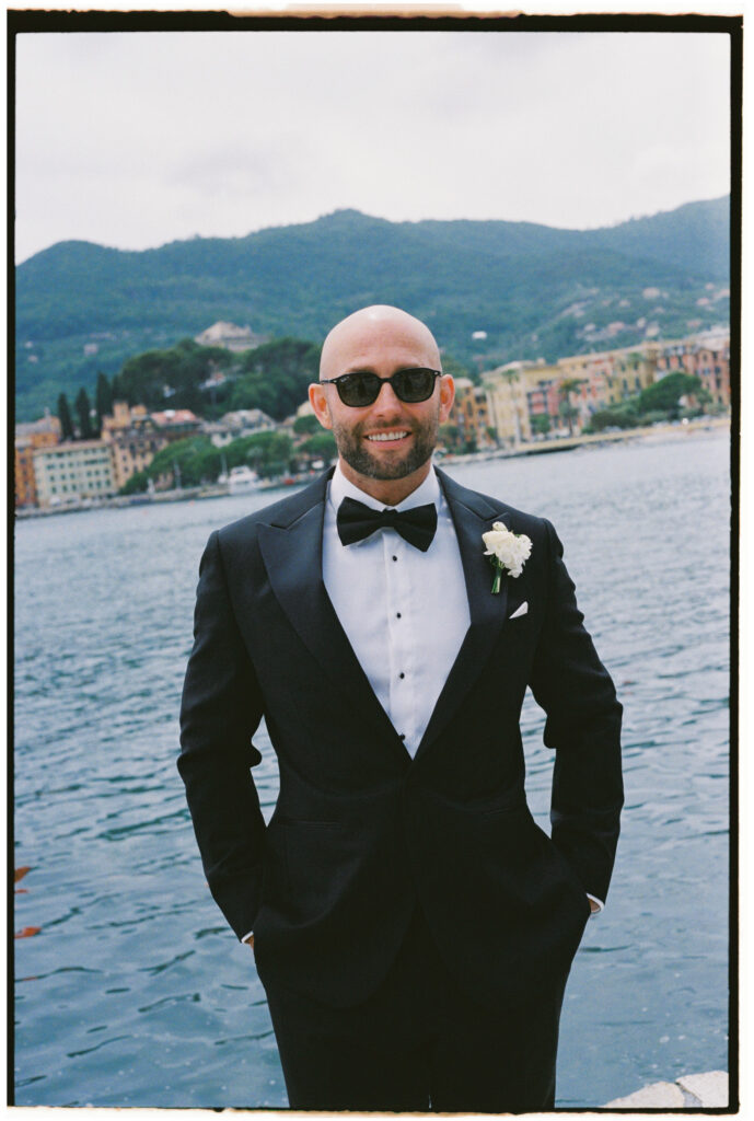 ilm photograph of groom in black tuxedo and sunglasses smiling by the water, Santa Margherita Ligure town visible across the bay