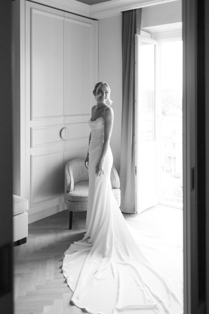 Black and white photo of bride in strapless wedding gown smiling by a window, viewed through an open doorway